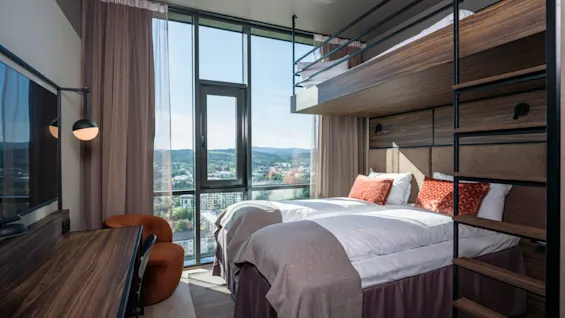 Et moderne hotelværelse med stor vinduesudsigt over byen, komfortable senge, en køjeseng og et skrivebord.