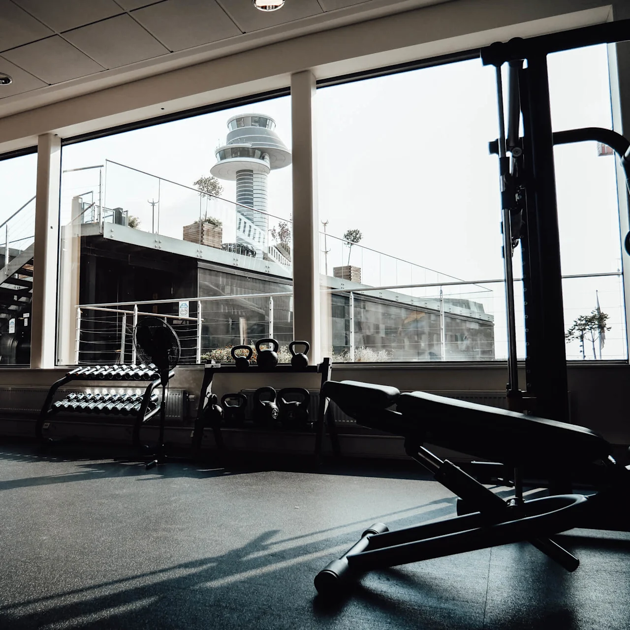 Exercise equipment is arranged in a gym with windows showing an outdoor area and a tall, round building. Rows of dumbbells and machines are situated on a rubber floor.