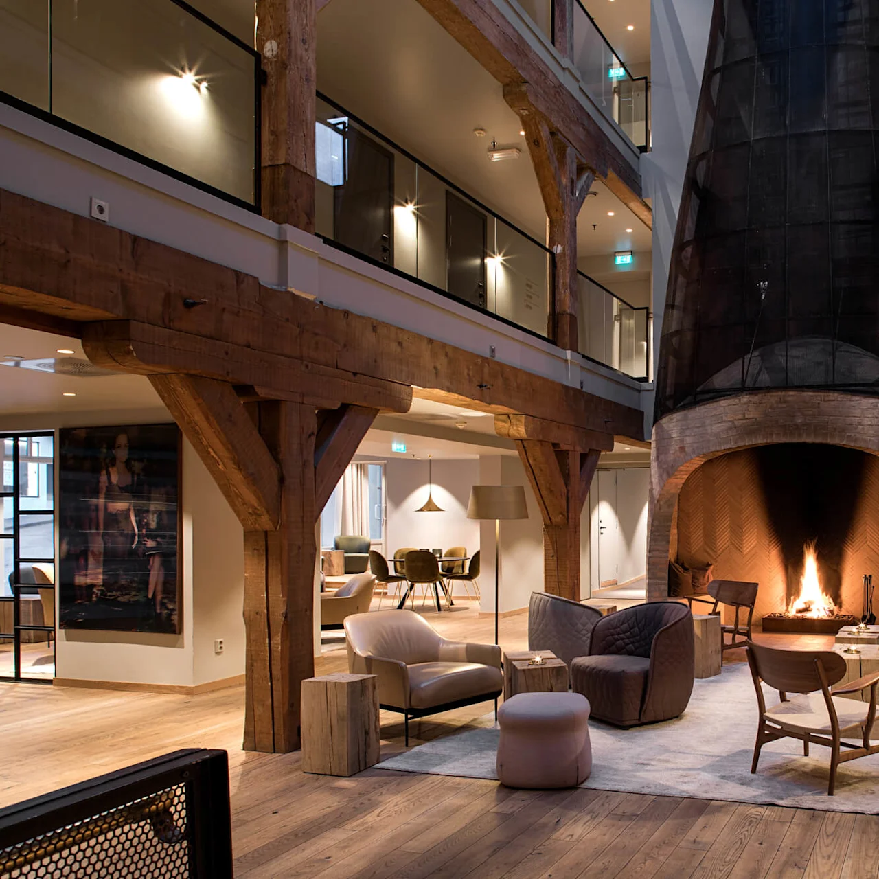 A fireplace crackles warmly, surrounded by modern seating in a rustic lounge. Timber beams accent the spacious two-story interior, filled with soft lighting and inviting decor.