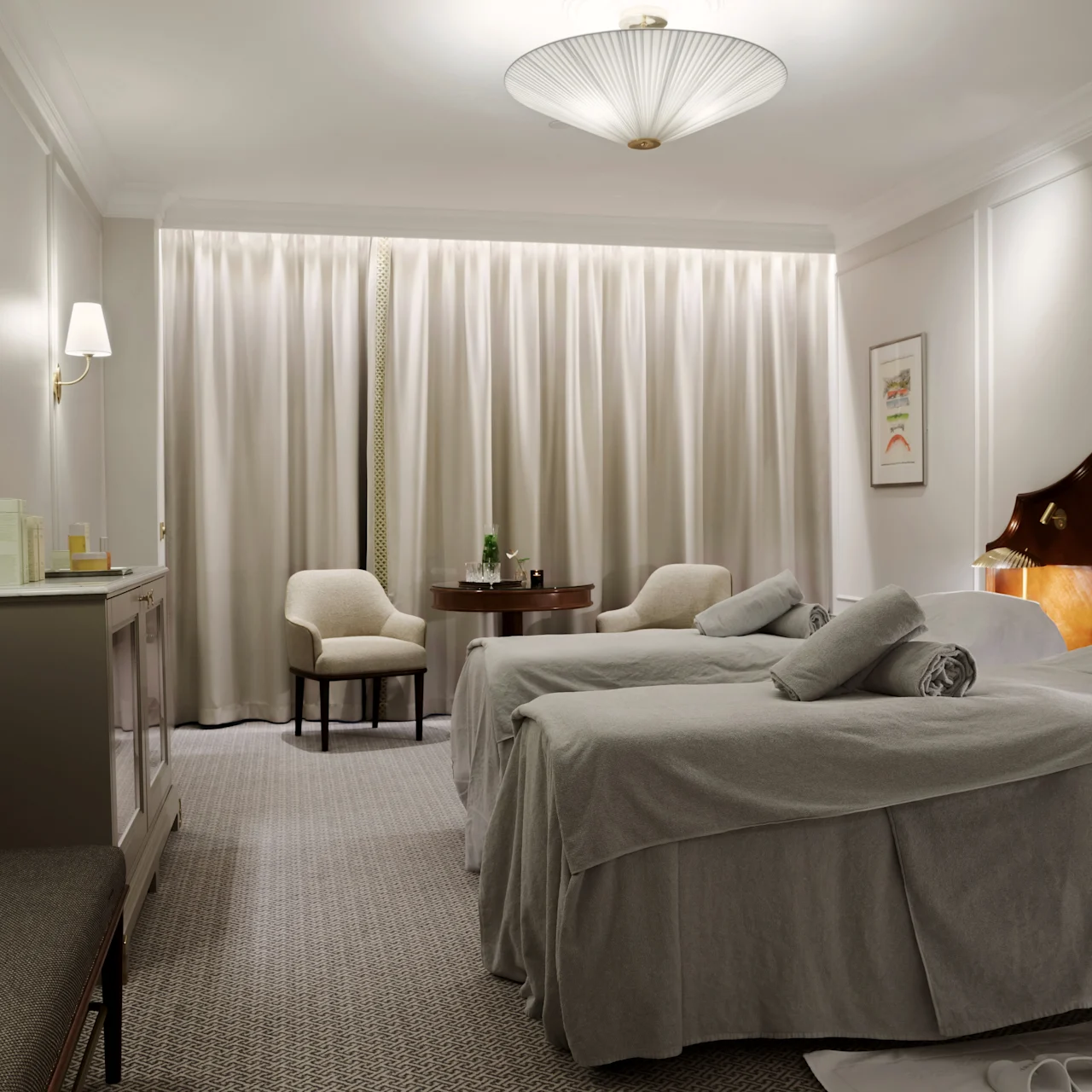 A hotel room features two neatly made beds with gray linens, bedside lamps glowing, a sitting area with chairs and a table near a window with drawn curtains.