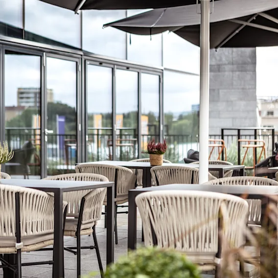 Utemøbler omgir bord, dekorert med potteplanter, på en moderne terrasse. Store glassvinduer i bygningen reflekterer omgivende grøntområder og bylandskap under delvis overskyet himmel.
