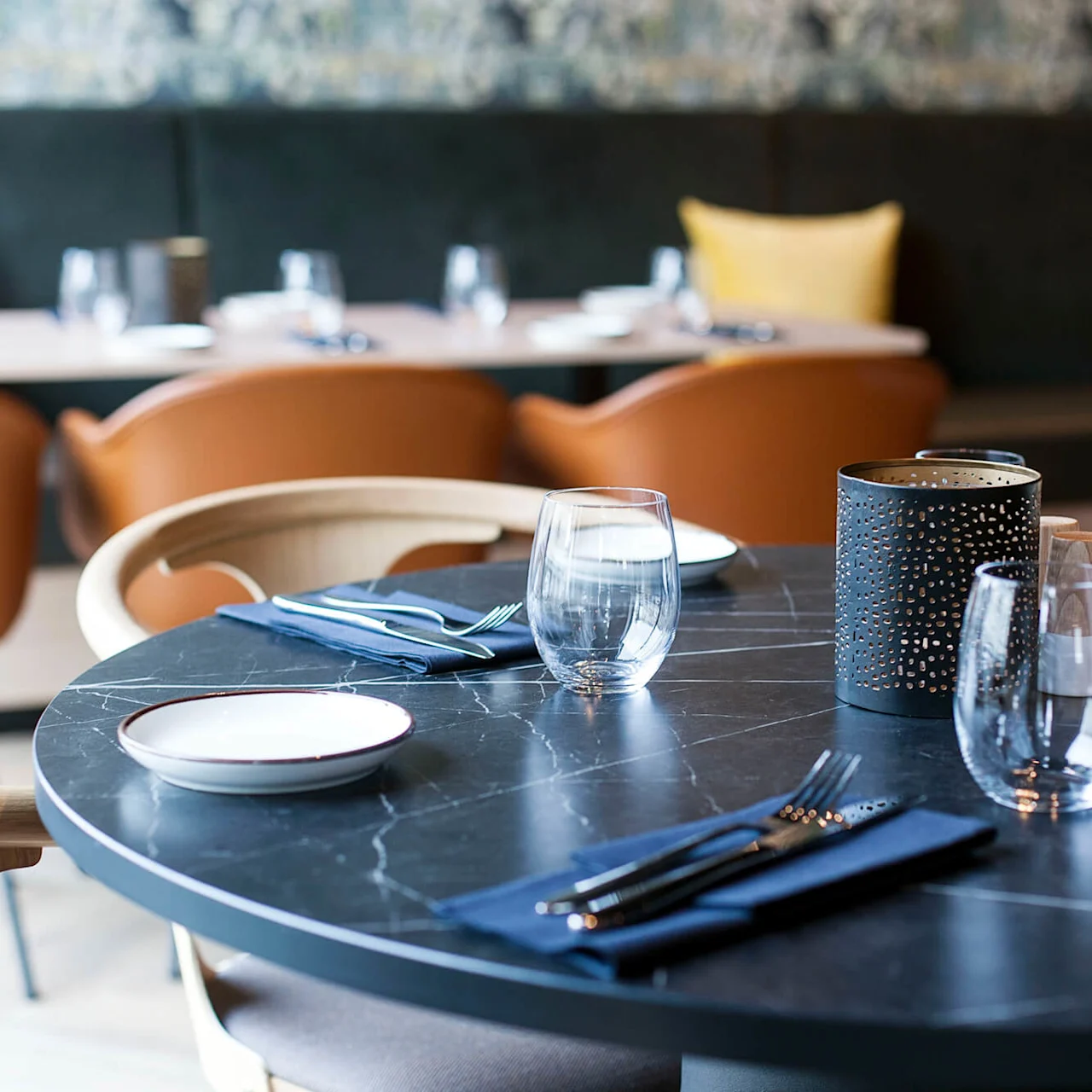 Et rundt svart marmorbord har elegant glass, bestikk og et midtstykke i en moderne, mykt opplyst restaurant med brune stoler i bakgrunnen.