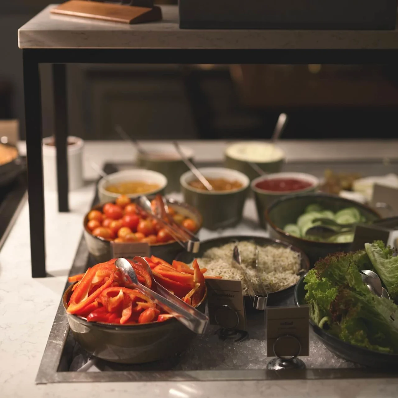 A buffet table with various condiments and fresh vegetables, labels indicating their names, under warm lighting.