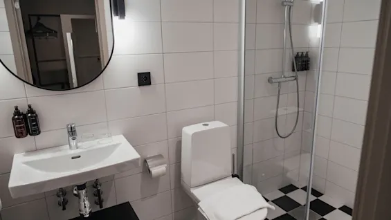 Ett stilrent badrum med vita kaklade väggar, ett modernt handfat, en toalett och en dusch med ett slående svartvitt rutigt golv.