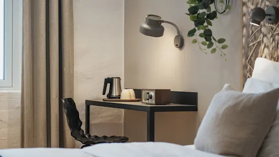 Viihtyisä hotellihuone, jossa sänky, työpöytä vedenkeittimellä ja kupeilla, seinävalaisin ja kasvi.