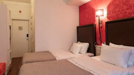 A cozy hotel room at Home Hotel Grand Bodø with two single beds, a red patterned accent wall, and a warm lamp. A door is visible on the left.