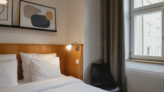 Viihtyisä hotellihuone siististi pedatulla sängyllä, modernilla lampulla ja abstraktilla taiteella seinällä. Home Hotel Uppsala.