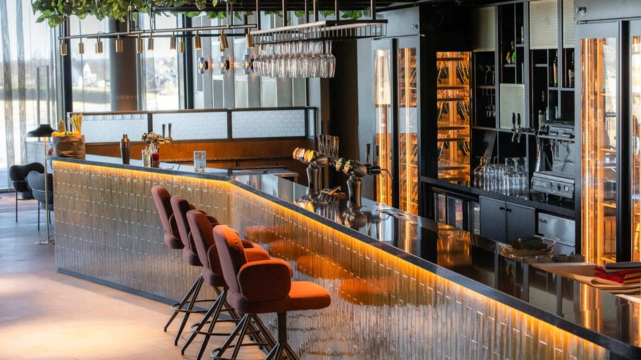En slank bardisk med pent arrangerte røde stoler og hengende glass. Varm belysning fremhever det moderne interiøret i en stilig, stemningsfull loungemiljø.