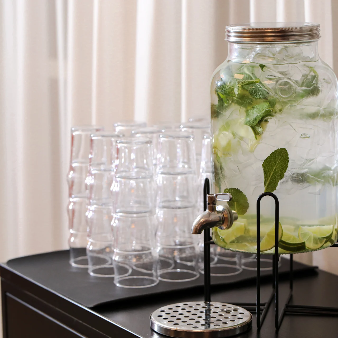A glass dispenser with cucumber, lemon, and mint-infused water rests on a stand, surrounded by stacked empty glasses on a black table against a backdrop of white curtains.