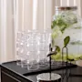 A glass dispenser with cucumber, lemon, and mint-infused water rests on a stand, surrounded by stacked empty glasses on a black table against a backdrop of white curtains.