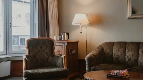 En mysig hotellrumslounge med fåtölj, soffa, soffbord med böcker och golvlampa, vilket skapar en avkopplande atmosfär.