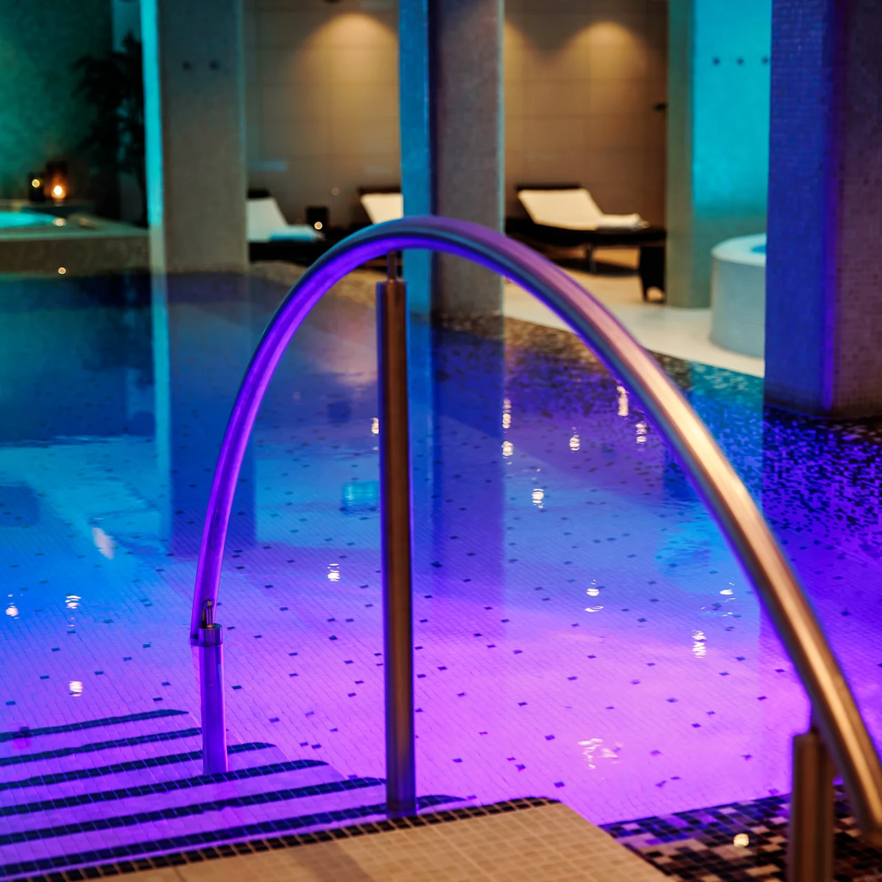 A swimming pool with underwater lights casts a blue and purple glow. Metal handrails descend into the water, surrounded by columns and lounge chairs, suggesting a luxurious indoor spa setting.