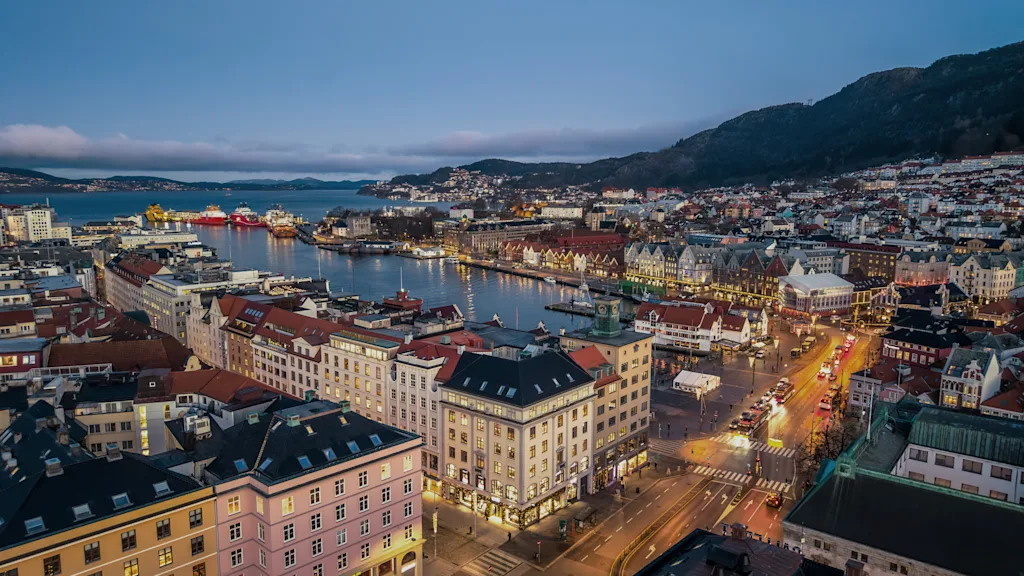 Bergen yöllä: Kaupungin valot valaisevat rakennuksia ja katuja vilkkaan sataman äärellä, vuoret taustalla.
