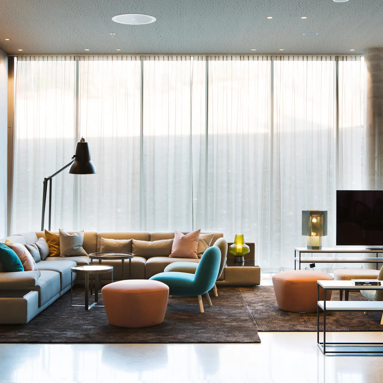 A modern living room features a large sectional sofa with colorful cushions, surrounded by lamps and a television. Floor-to-ceiling windows are veiled with sheer curtains.