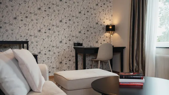 A cozy room with floral wallpaper, a desk and chair, a sofa, and a coffee table with books, suggesting a comfortable space for relaxation or work.