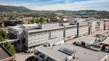 Et stort hotell merket "Quality Airport Hotel" står i et urbant område, omgitt av bygninger, veier og trær, med fjerne åser under en blå himmel.
