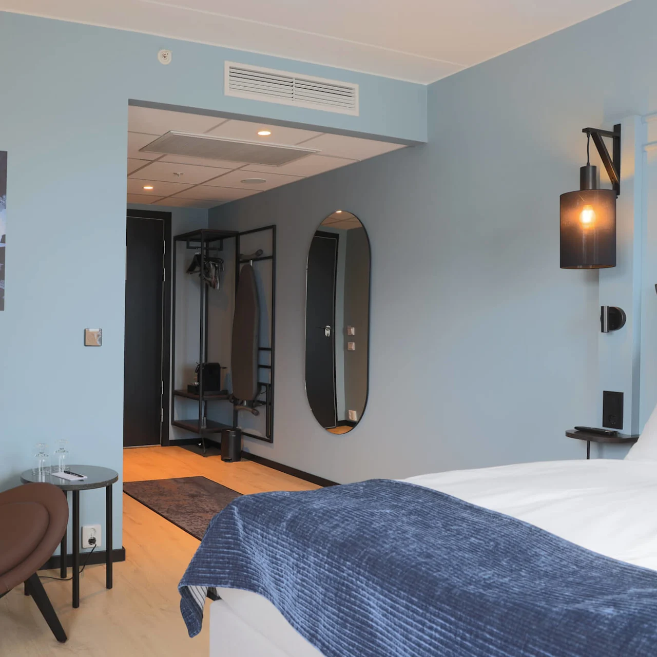 A modern hotel room with a brown chair, bed, nightstands, and wall decor. The ambiance is cozy with muted lighting.
