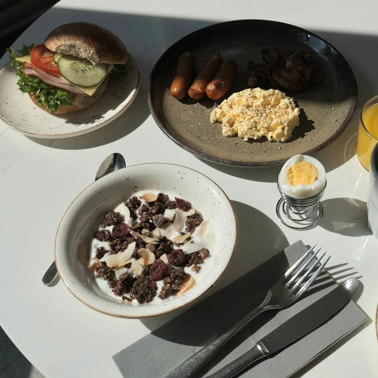 En morgenmad med en sandwich med salat, tomat, agurk, røræg, pølser, yoghurt med granola, kogt æg, kaffe, juice og frugtskiver på et hvidt bord.