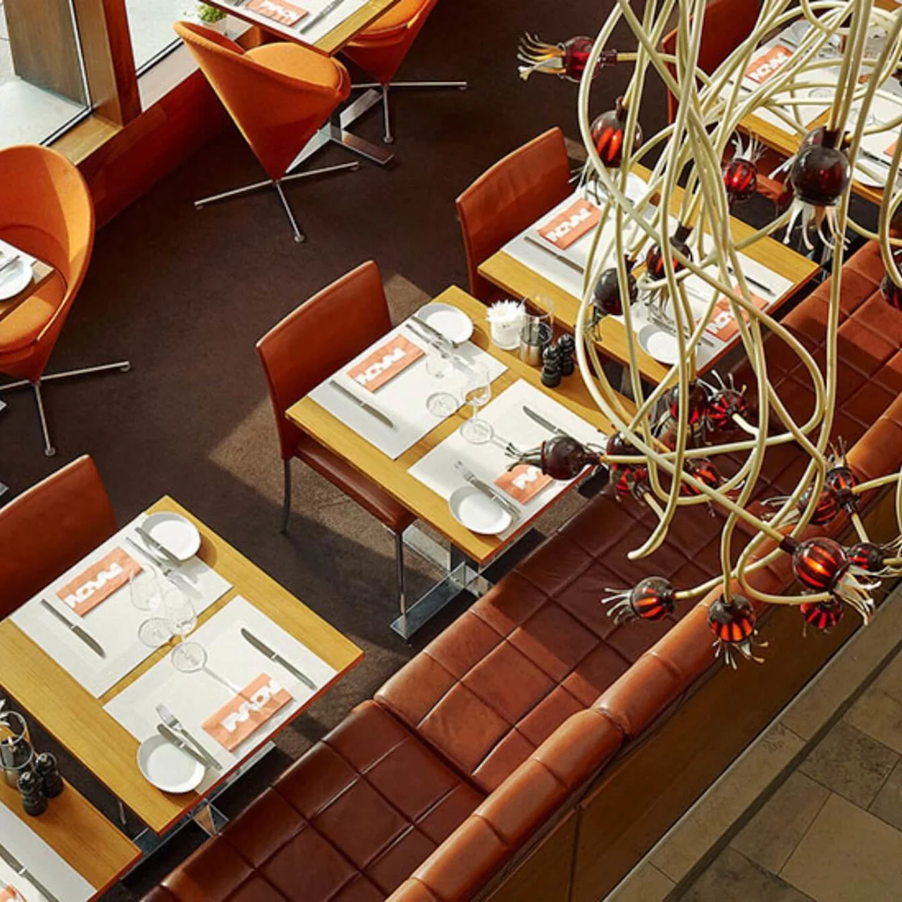 Restaurant tables are set for dining with white plates, glasses, and utensils on light wooden surfaces. Orange chairs and a decorative overhead chandelier create a stylish and inviting atmosphere. Napkins read "NOMA."