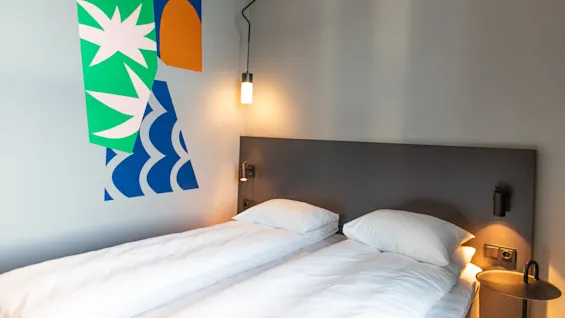 Two neatly-made twin beds rest side by side against a wall; abstract art decorates the wall above, and small lamps provide light, creating a cozy atmosphere.
