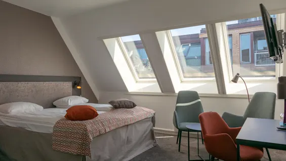 Et pænt hotelværelse har en seng med puder og et dekorativt tæppe. Sollys strømmer igennem skråvinduer og oplyser to stole og et lille bord ved siden af et vægmonteret TV.