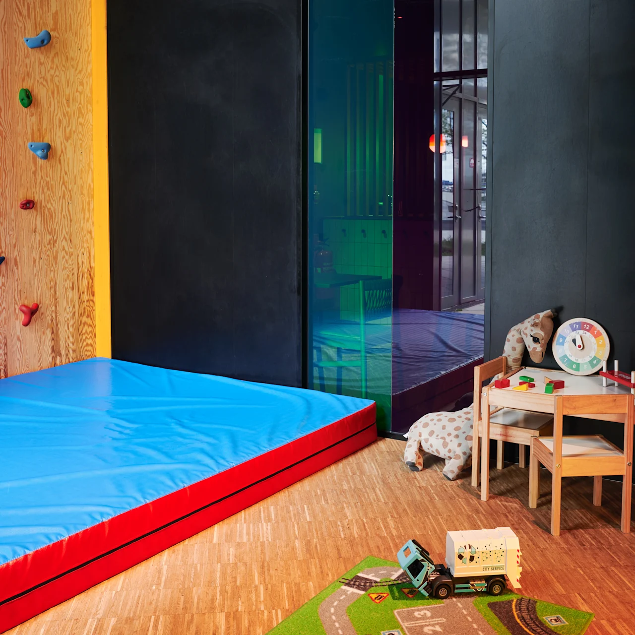 A children's playroom features a climbing wall with colorful holds and a soft blue mat. Adjacent, a small table holds toys and a chair. Nearby, a rug displays roads.