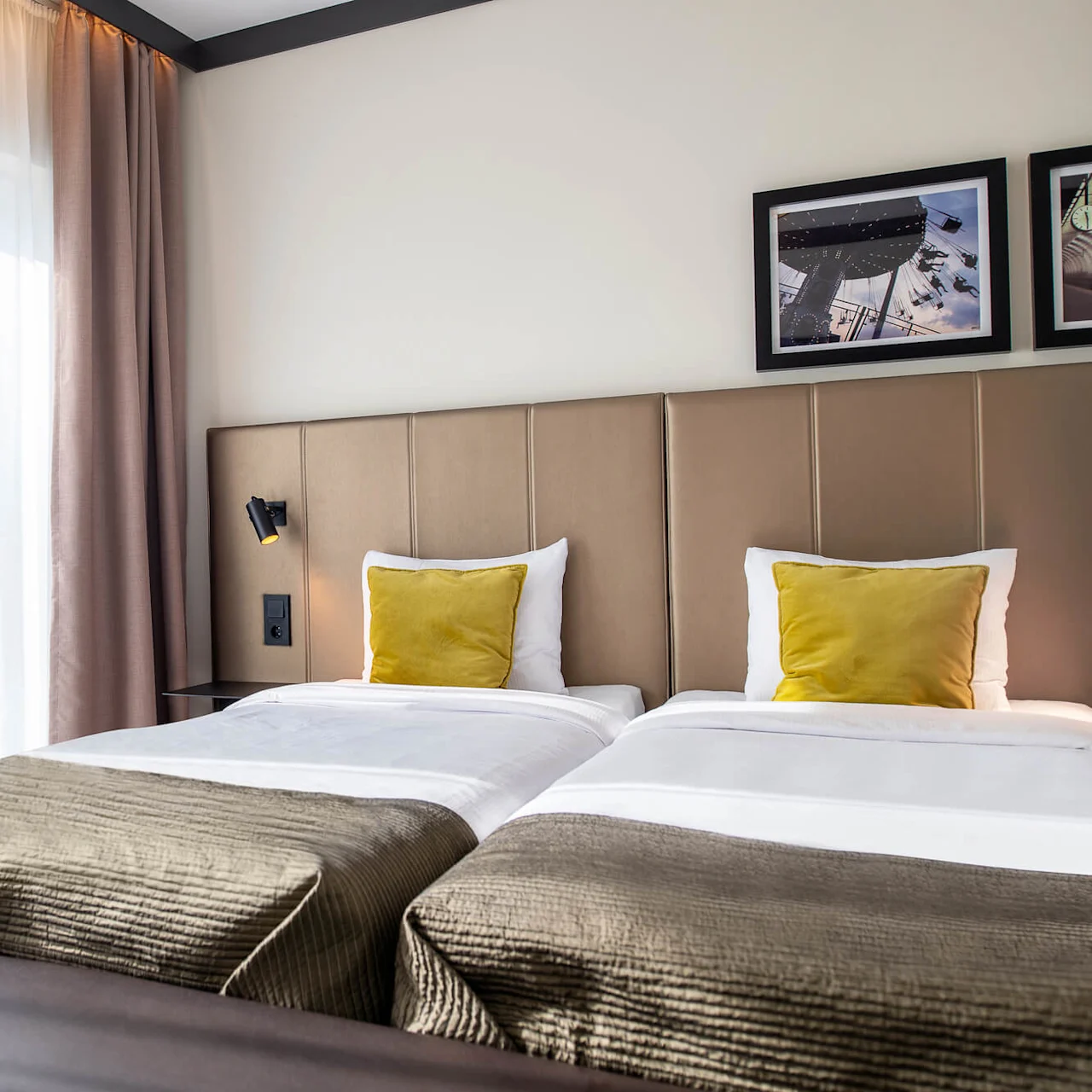Two neatly made twin beds with yellow cushions, under framed artwork, situated in a warmly lit room with beige curtains covering large windows.