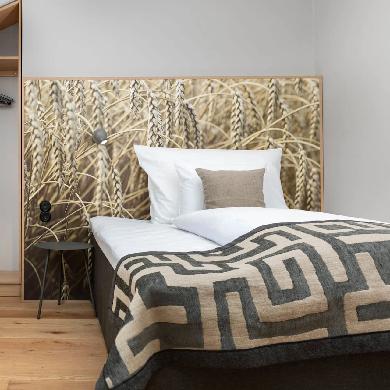 A well-lit, tidy hotel room with wooden floors, a plush bed, patterned headboard, a cozy chair, pillows, a standing lamp, and built-in shelves.