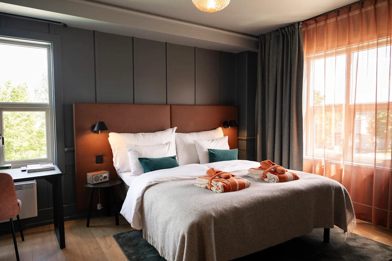 En säng med orange handdukar och gröna kuddar står i ett modernt hotellrum med ett skrivbord, stora fönster och mjuk belysning.
