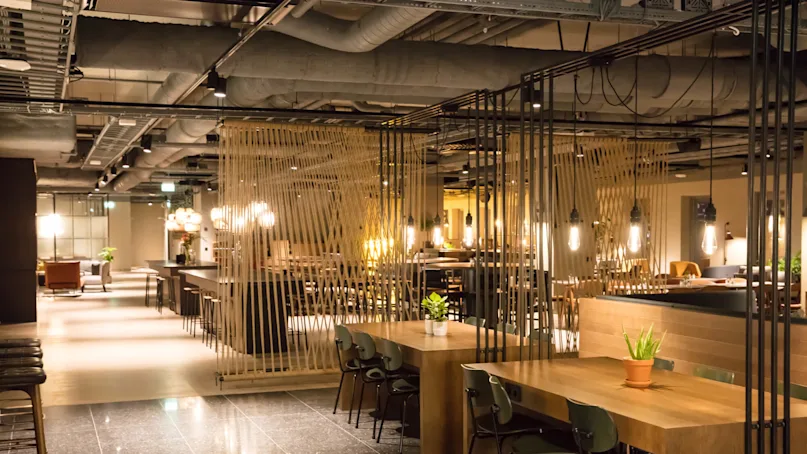 A modern restaurant with warm lighting, wood furniture, and hanging rope dividers. Exposed ceiling pipes contribute to an industrial vibe.