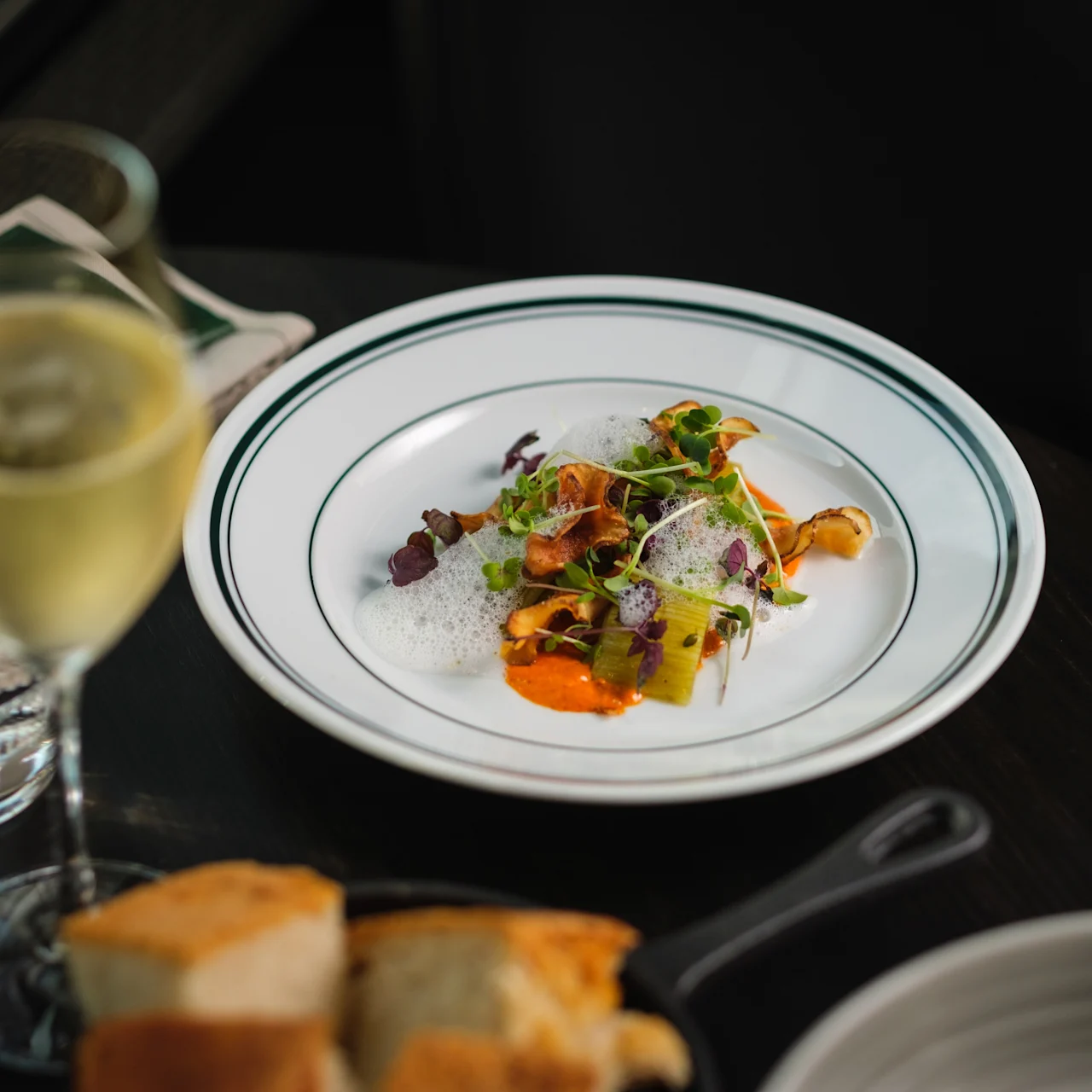 En gourmeträtt med färgglada grönsaker och skum ligger elegant på en vit tallrik, åtföljd av ett glas vitt vin och bröd, placerad på ett mörkt bord.