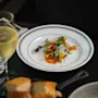 En gourmeträtt med färgglada grönsaker och skum ligger elegant på en vit tallrik, åtföljd av ett glas vitt vin och bröd, placerad på ett mörkt bord.