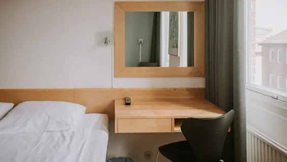Et skrivebord af træ med en fjernbetjening ligger under et vægmonteret spejl. En seng med hvidt sengetøj er ved siden af, og et vindue med gardiner er til højre.