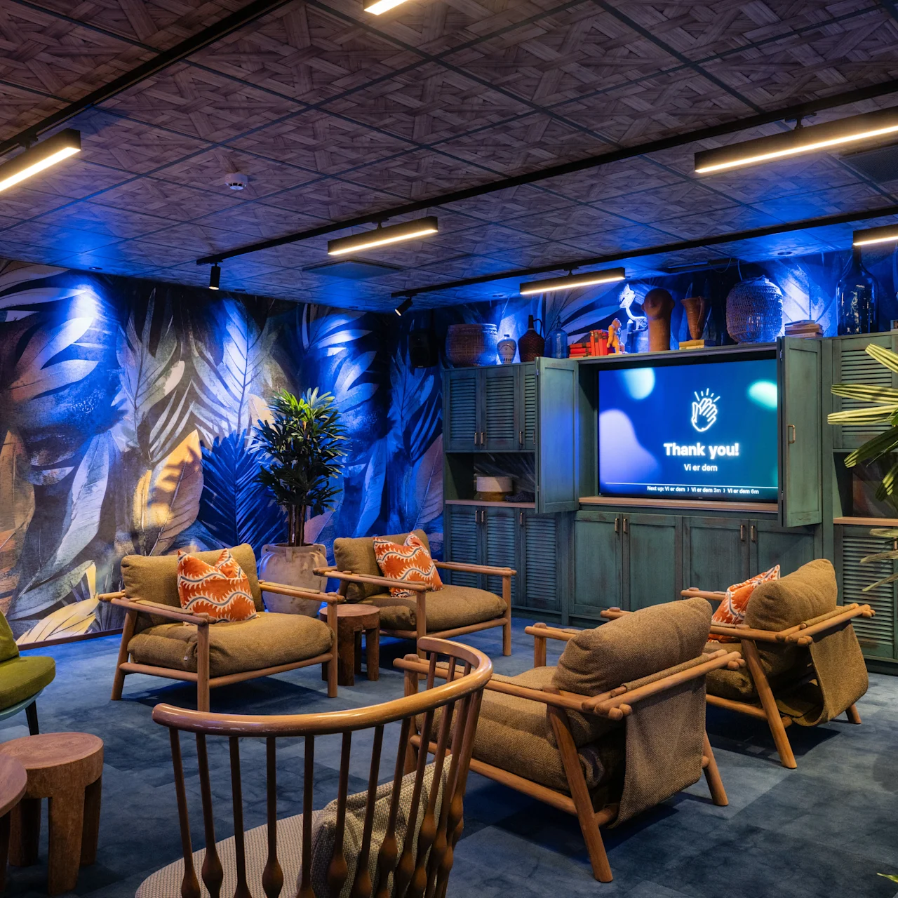 Cozy lounge with wooden chairs and green cushions facing a screen displaying “Thank you! Merci beaucoup! Gracias!” against blue-lit tropical leaf wallpaper, surrounded by plants and soft lighting.