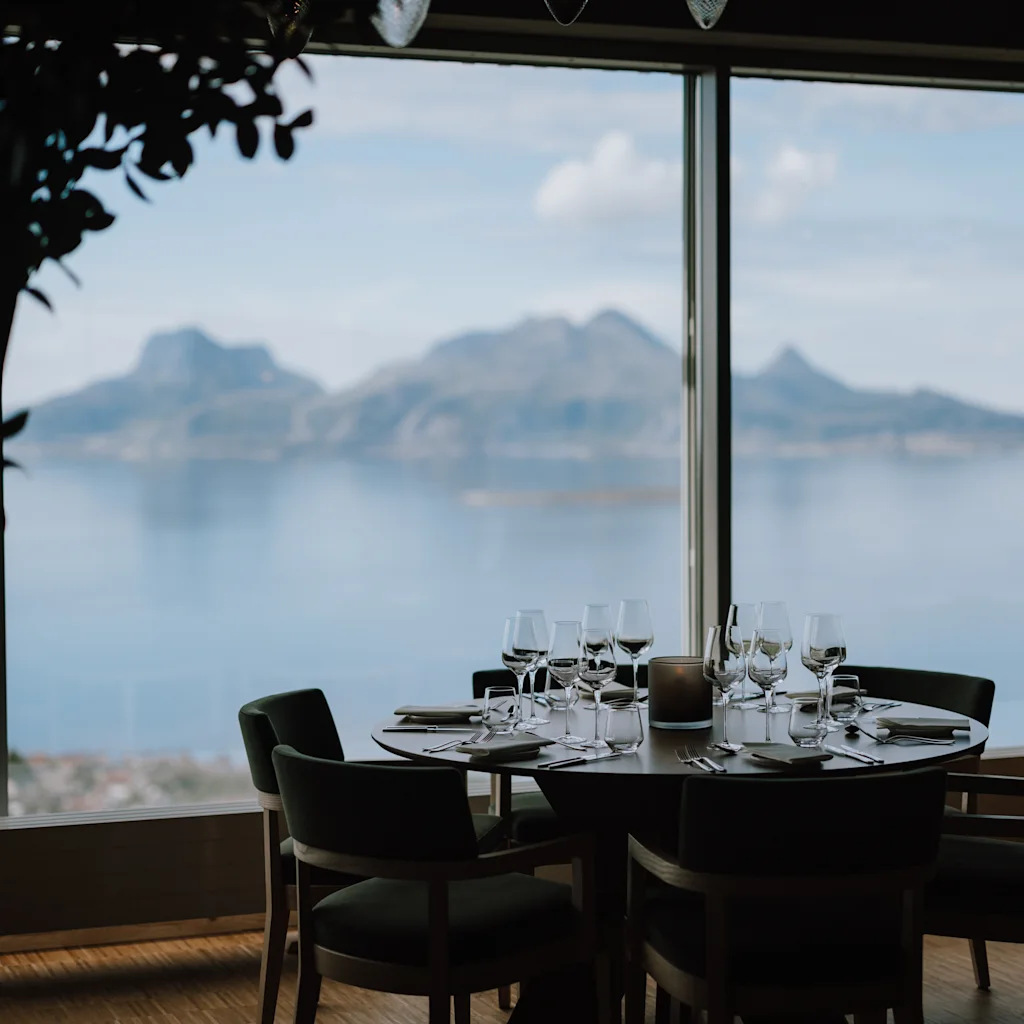 Ett bord med snyggt arrangerade glas och bestick har panoramautsikt över berg och vatten genom ett stort fönster. En person står i närheten i en restaurangmiljö.