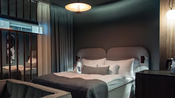 En säng med vit och grå bäddning är prydligt arrangerad, belägen i ett svagt belyst, modernt sovrum med reflekterande ytor, gardiner och en mysig atmosfär.
