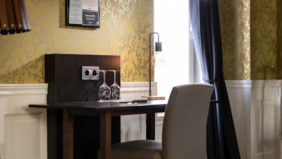 A wooden desk with a chair and lamp sits by patterned wallpaper. Two upside-down glasses and electrical outlets are on the desk beneath a framed notice.