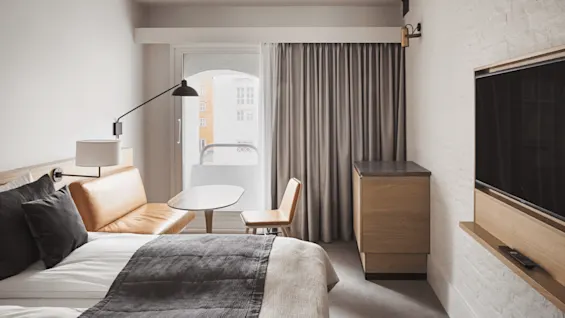 A modern hotel room features a bed with gray and white bedding, a leather sofa, and a small table. A large window with curtains provides natural light.