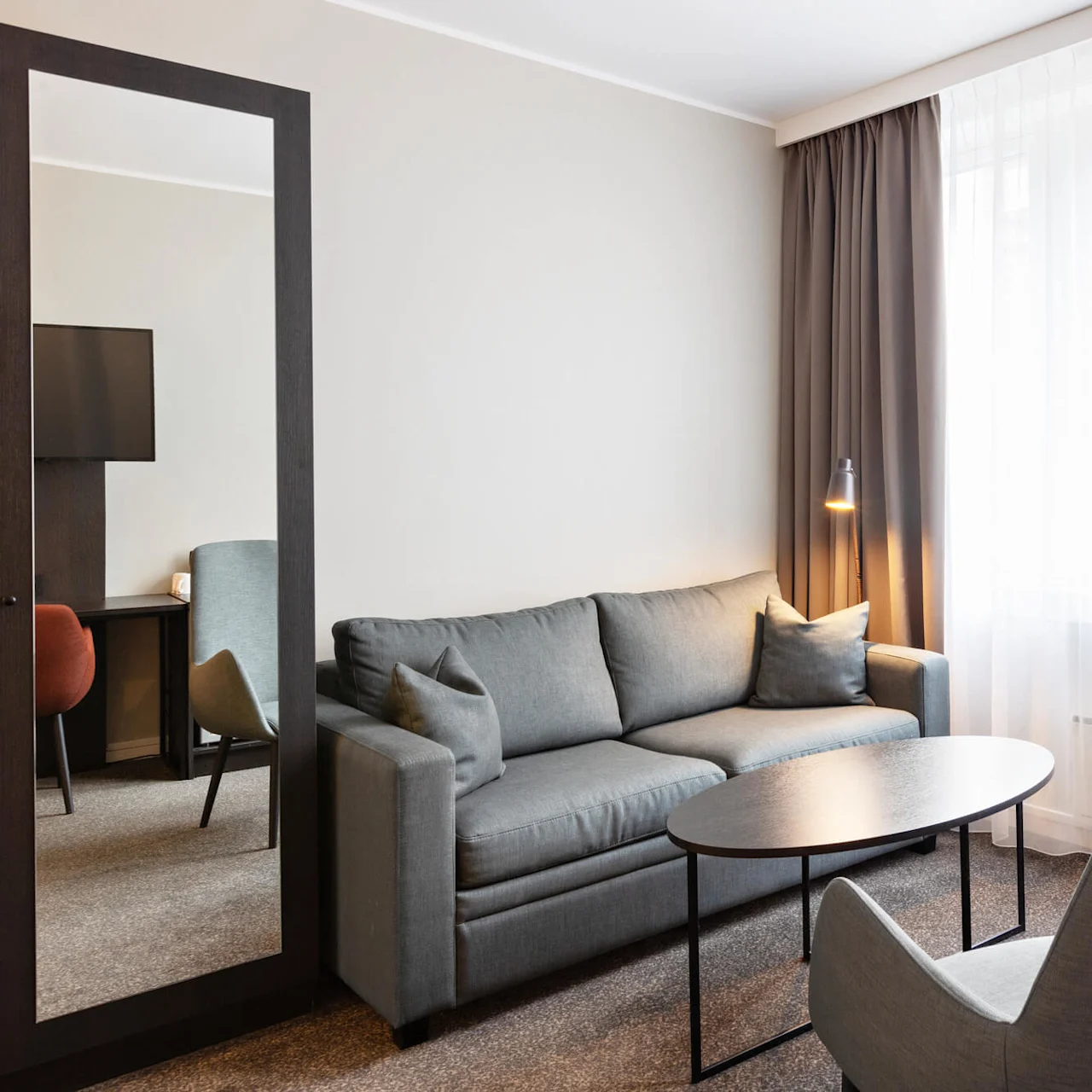 En grå sofa står i stuen med to stole og et sort sofabord. En gulvlampe oplyser rummet ved siden af ​​lukkede gardiner, med et vægspejl, der reflekterer en del af et skrivebord.