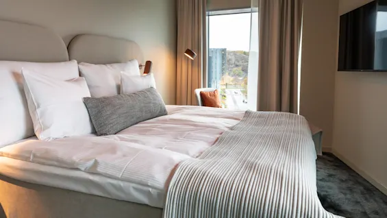 En prydligt bäddad säng, prydd med vita och grå kuddar, står bredvid ett fönster med gardiner i ett hotellrum. En väggmonterad TV och stadsutsikt kompletterar miljön.