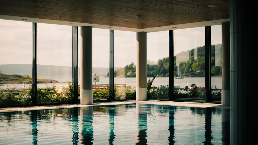 En indendørs swimmingpool med store vinduer, der tilbyder en naturskøn udsigt over en sø omgivet af frodige grønne bakker under en lys himmel.