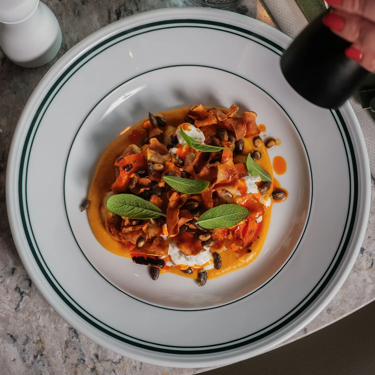 A plate of colorful pasta with vegetables and herbs is being seasoned with pepper. It's placed on a marble table with green napkins and an S-shaped logo.