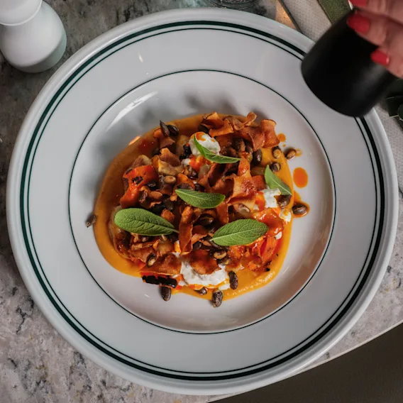 En tallerken med farverig pasta og grøntsager bliver krydret med peber. Den står på et marmorbord med grønne servietter og en S-formet logo.