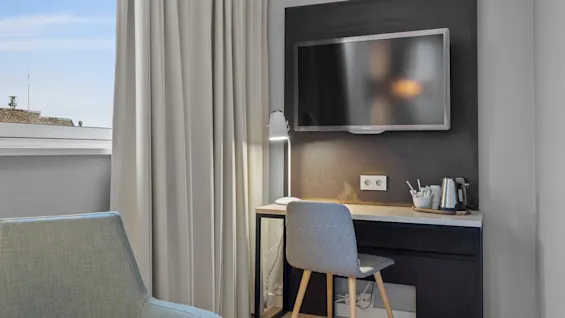 Ett mysigt hotellrum med en väl upplyst skrivbordsplats med TV, en bekväm stol och ett fönster med naturligt ljus.