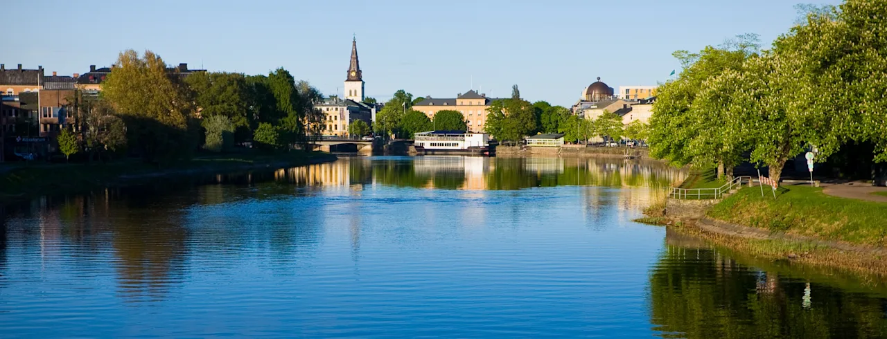 Rolig flod, der afspejler den klare blå himmel omgivet af frodige grønne træer og bygninger i baggrunden, herunder et fremtrædende kyrkespir under en solrig dag.