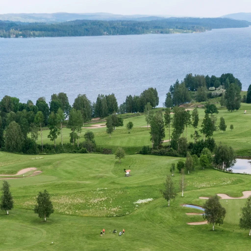 En vidsträckt grön golfbana med spridda träd och sandfällor, mot bakgrund av en bred, lugn sjö.