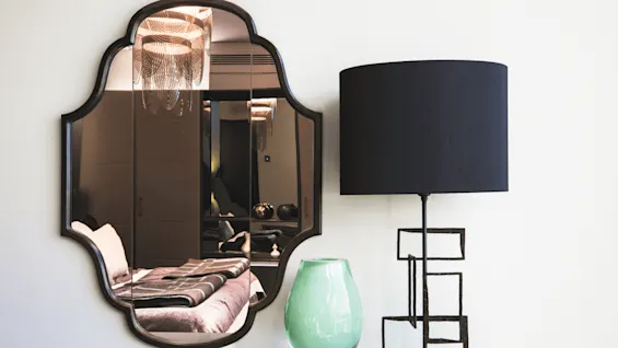 Et dekorativt speil reflekterer en lysekrone og en seng i et rom. En svart lampe med geometrisk base og en grønn vase står på den tilstøtende overflaten.