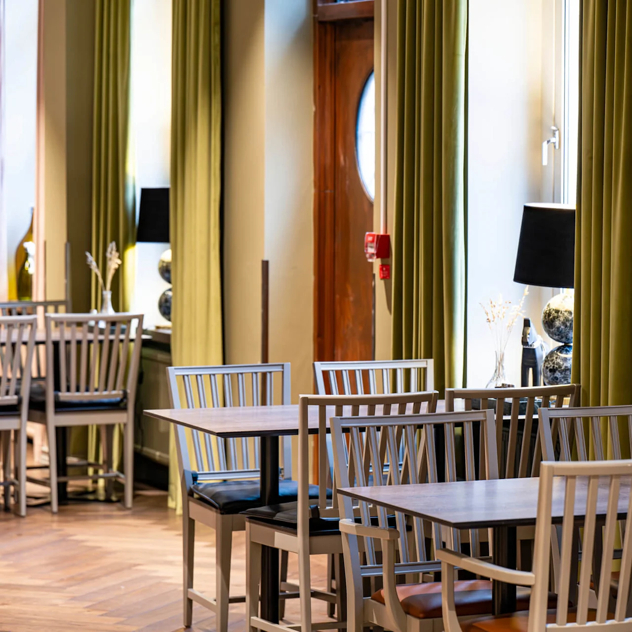 Wooden tables and chairs set up neatly in an empty modern dining room with large windows, curtains, and decorative lamps.