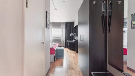 A hotel room hallway leading to a bed, desk, and window. A dark wardrobe with a mirror is on the right.