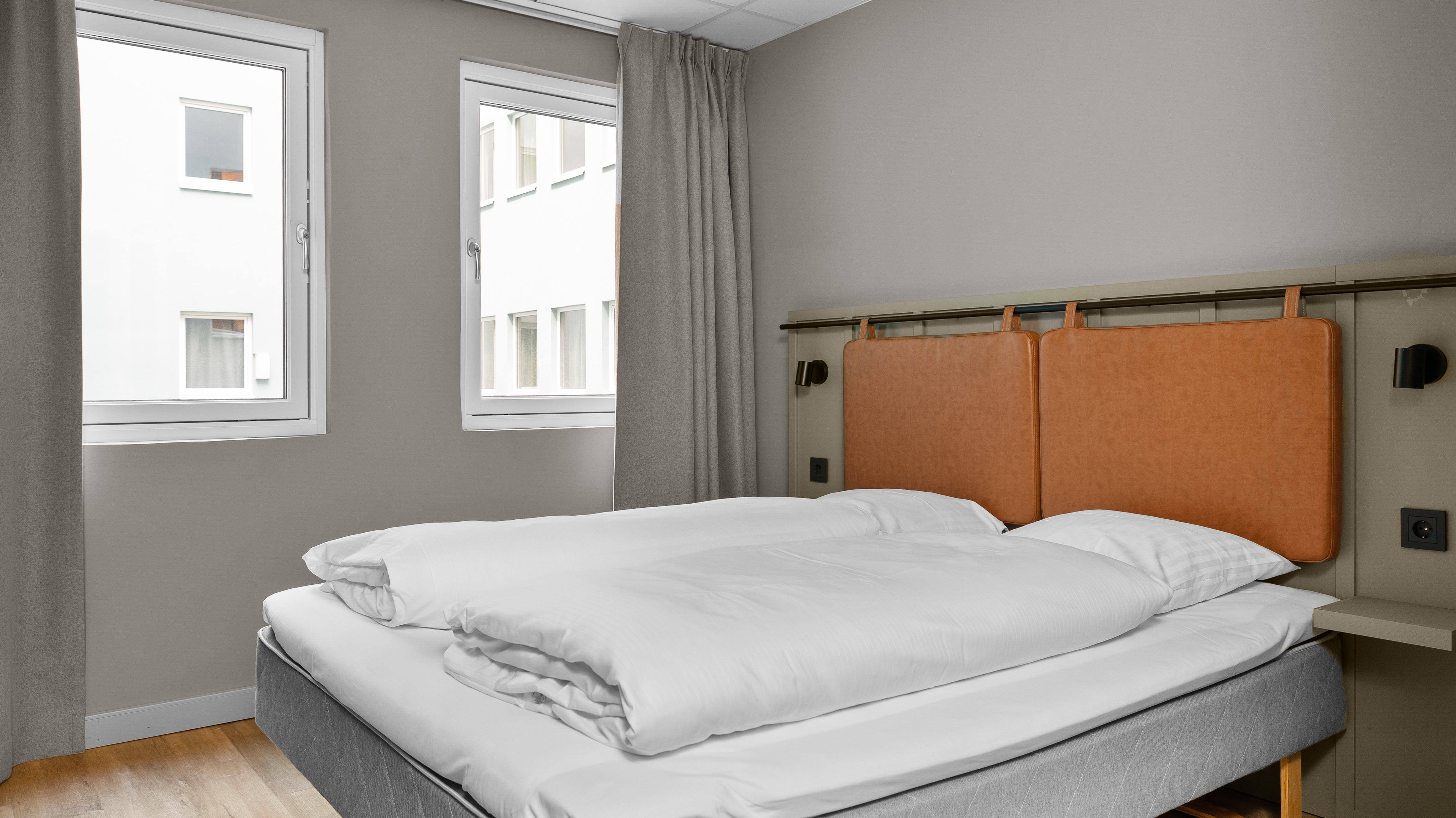 Et hyggeligt hotelværelse med en komfortabel seng, hvidt sengetøj og to vinduer, der slipper naturligt lys ind. Perfekt til et afslappende ophold.